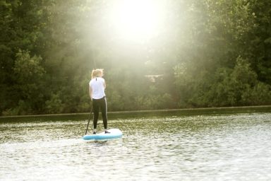 Frau auf einem Stand-Up-Paddle-Board auf einem ruhigen Gewässer, Sonnenlicht im Hintergrund.