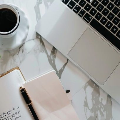Schreibtisch mit Notizbuch, Stift, Laptop und einer Tasse Kaffee.